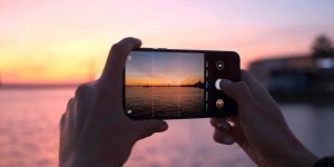 A pair of hands, presumably belonging to a person (not pictured) hold a smartphone in landscape mode, taking a picture of a sunset over the water.