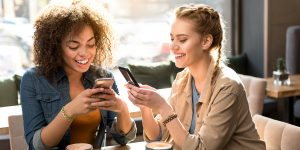 Two women, having coffee, smiling while on their mobile phones.