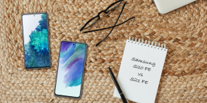 A Samsung Galaxy S21 FE 5G and Samsung Galaxy S20 FE 5G on a mat next to a pair of glasses and a notepad that has both phone names written on them