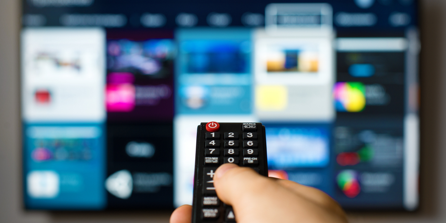 A hand pointing a remote control at an out-of-focus television screen displaying a variety of streaming apps.