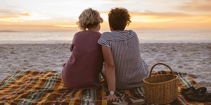 A couple share the view of a sunset from a beach blanket.