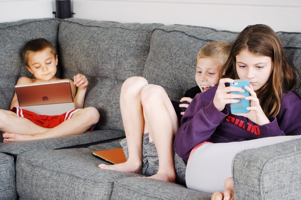 Three children playing on devices while sitting on a sofa together. 